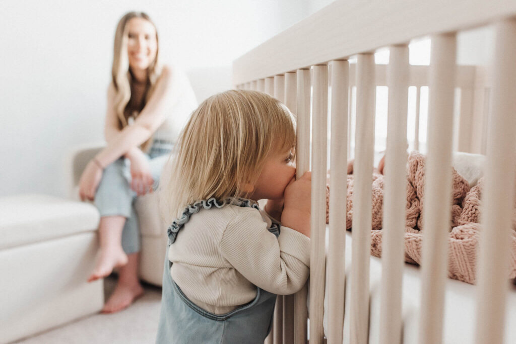 A toddler girl peeks through her newborn sisters crib in their frisco tx home